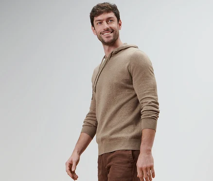 Ein lächelnder Mann trägt einen Strick-Hoodie und braune Hosen.