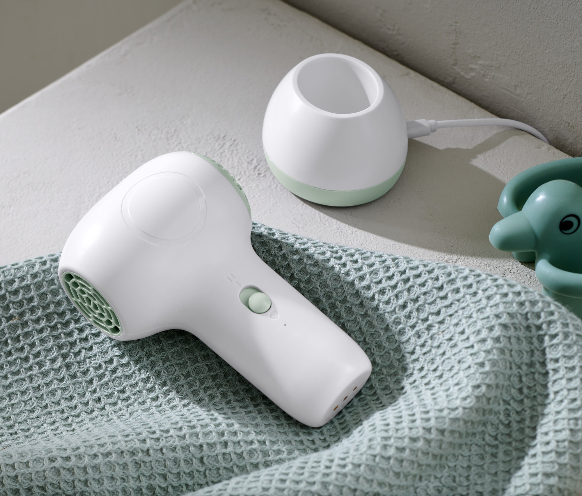 Ein weißer Akku-Baby-Haartrockner liegt auf einem hellblauen Handtuch neben einer Ladestation und einem Badespielzeug.