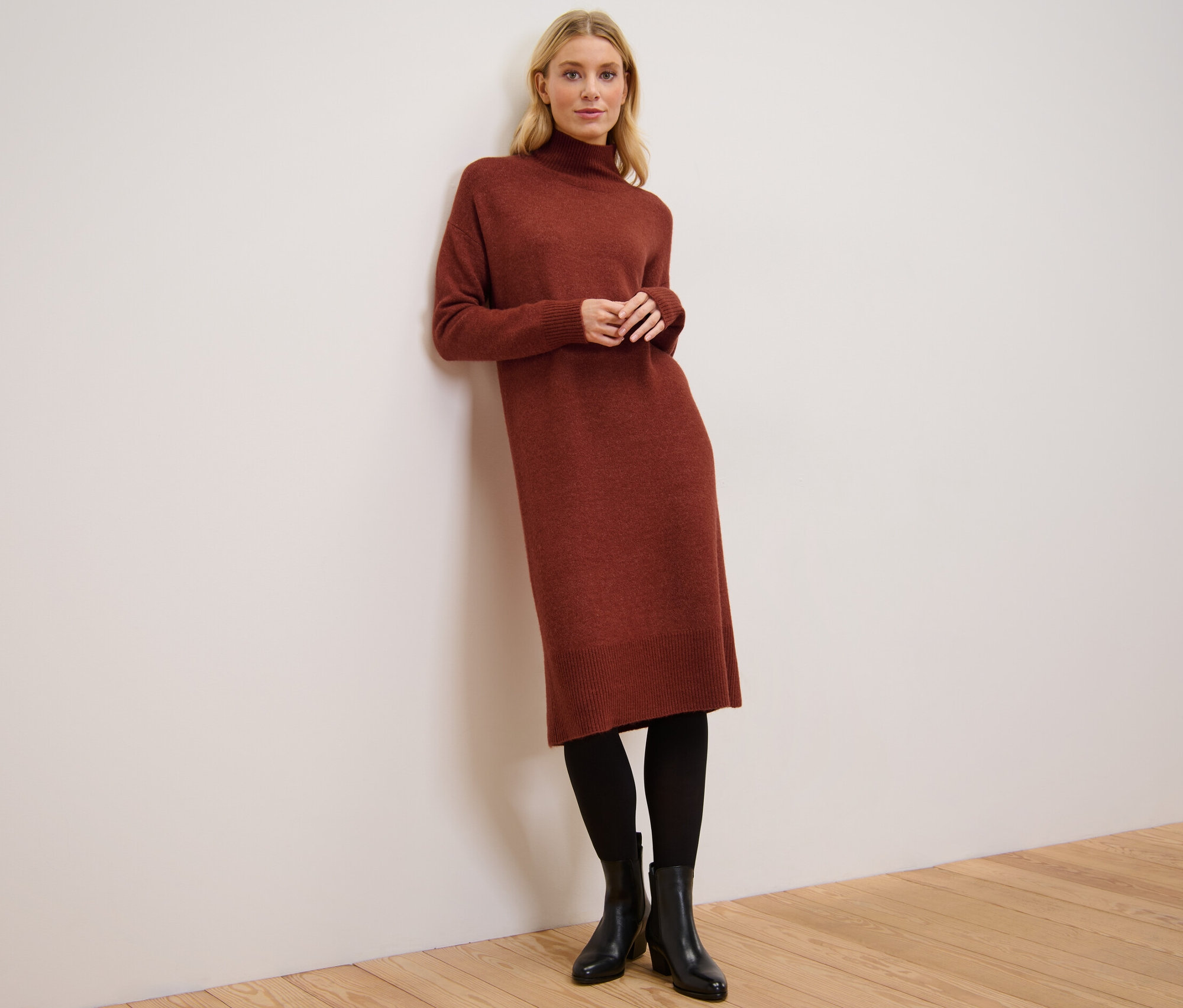 Frau lehnt an Wand, trägt Strickkleid mit Wollanteil, schwarze Strumpfhose und Stiefeletten.