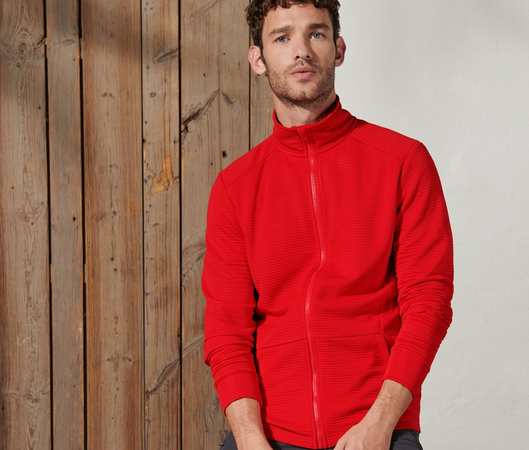 Mann sitzt auf Holzbalken und trägt rotorange Funktionsjacke.