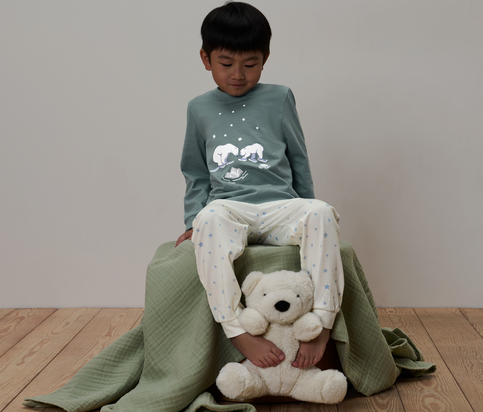 Junge sitzt auf grüner Decke und trägt einen grünen Kinder-Pyjama »Glow in the dark« mit Bären-Motiv.