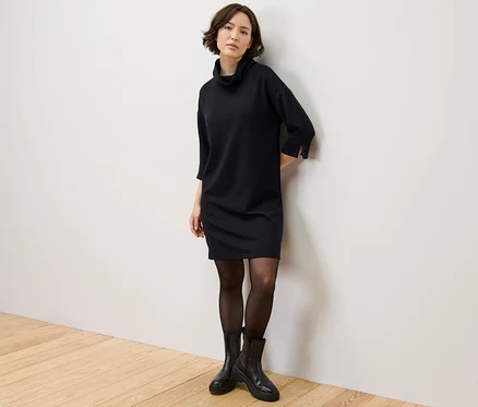 Frau steht an der Wand und trägt ein Sweatkleid aus Modal-Mix, eine schwarze Strumpfhose und schwarze Stiefel.