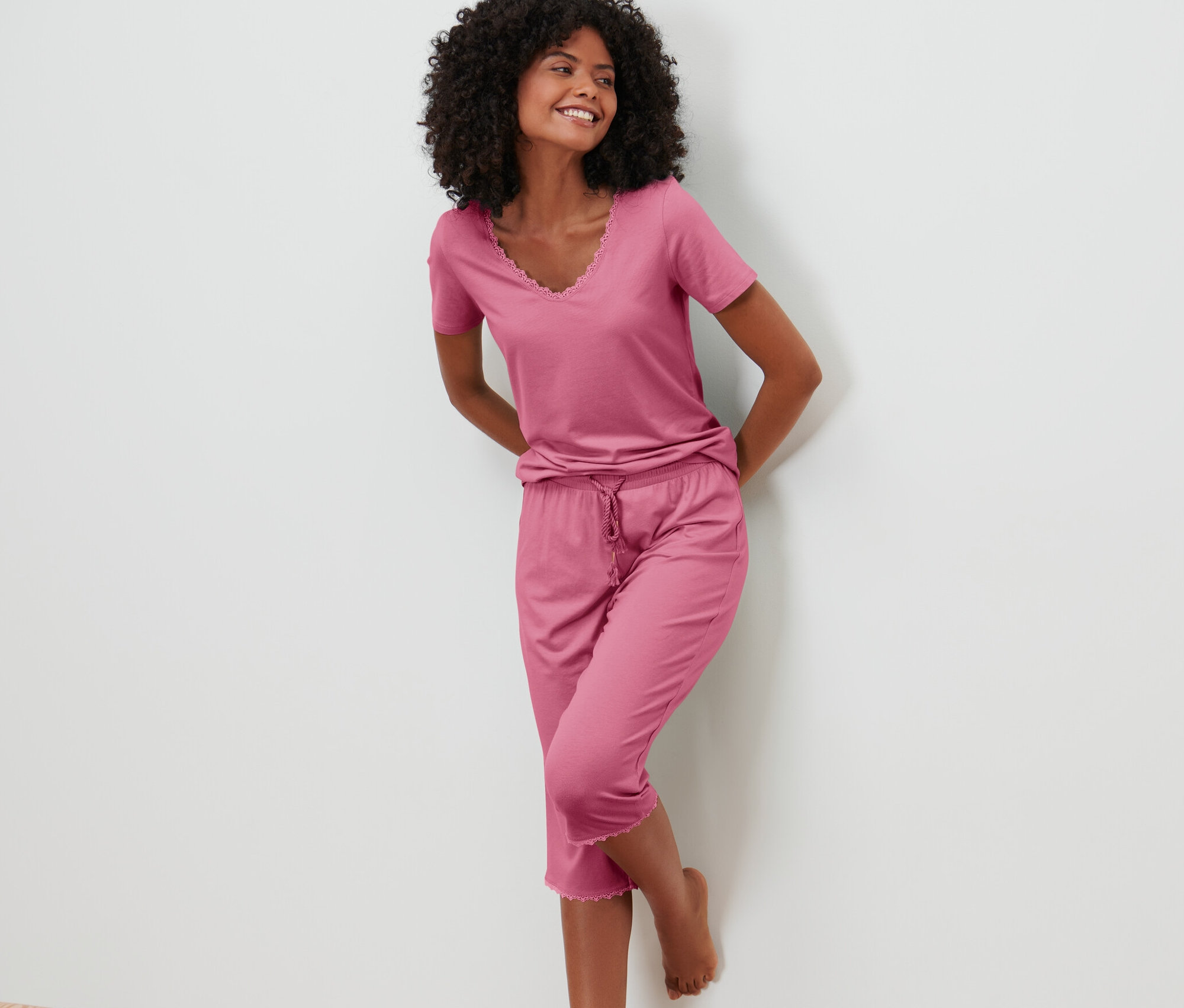 Lächelnde Frau lehnt an einer Wand und trägt ein rosafarbenes 3/4-Pyjama-Set.