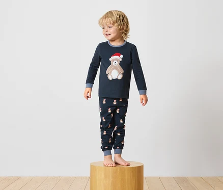 Junge steht auf einem Holzsockel und trägt einen dunkelblauen Pyjama mit einem Teddybär-Motiv.