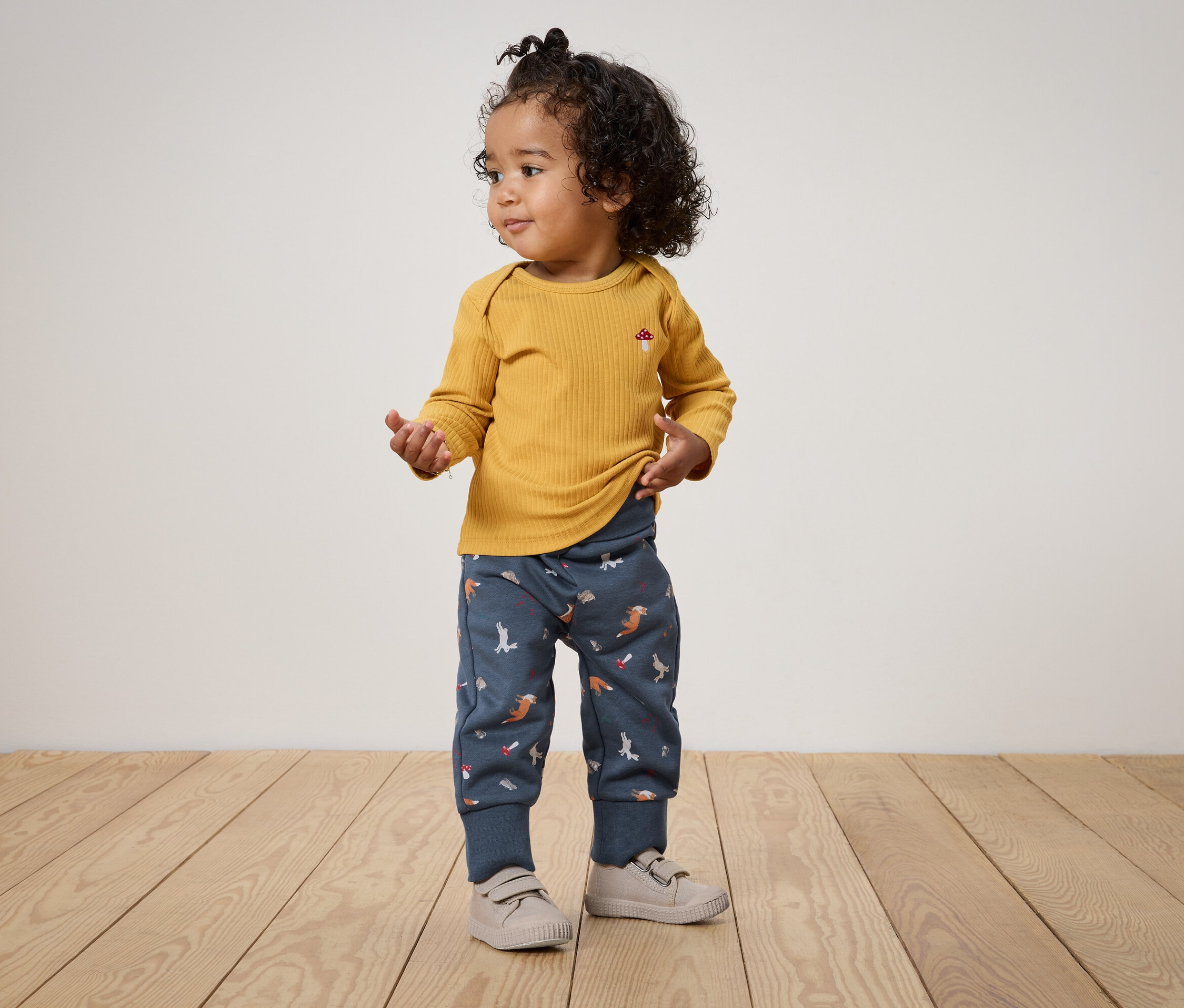 Ein Kind mit lockigem Haar steht auf einem Holzboden und trägt ein senfgelbes Langarmshirt und blaue Hosen mit Tiermotiven.