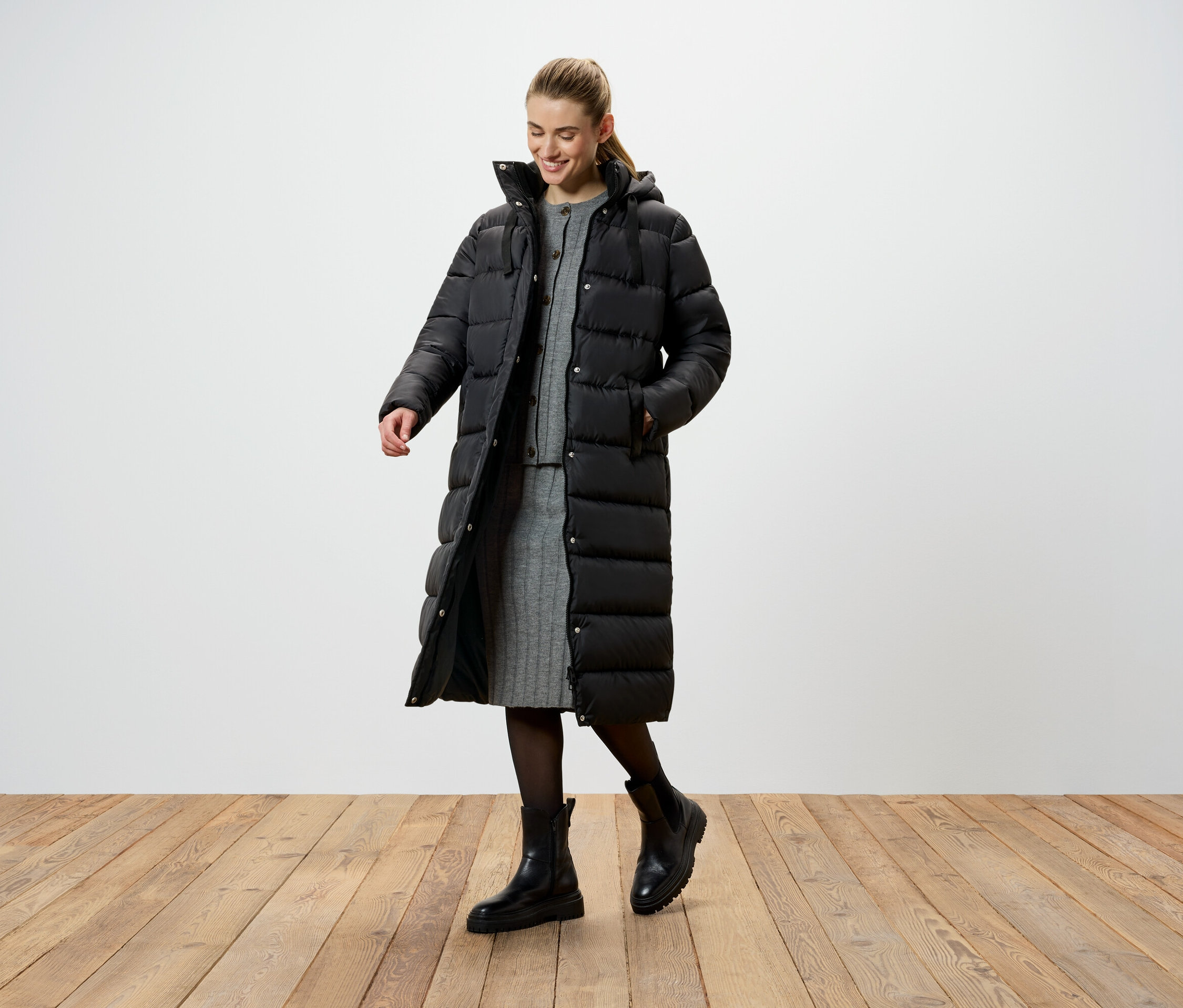 Eine Frau in einer langen, schwarzen Daunenjacke, einem grauen Strickkleid und schwarzen Stiefeln steht auf einem Holzboden.
