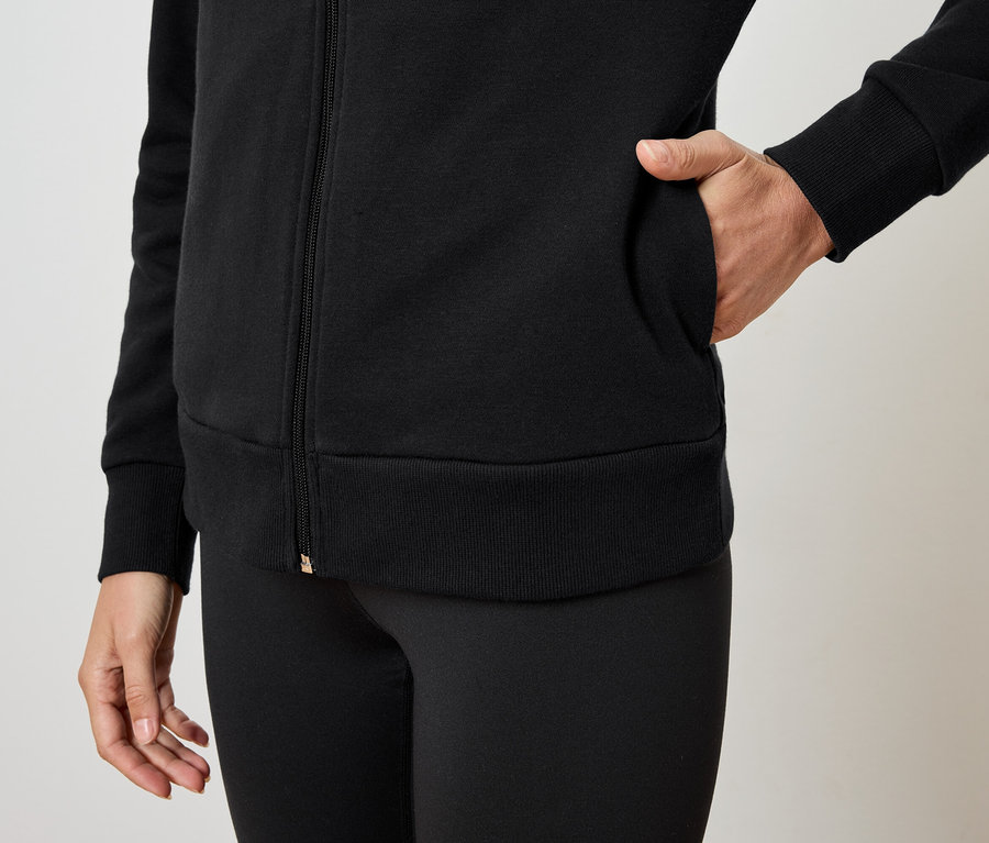 Frau hat die Hand in der Tasche einer schwarzen Reißverschlussjacke und trägt schwarze Leggings.
