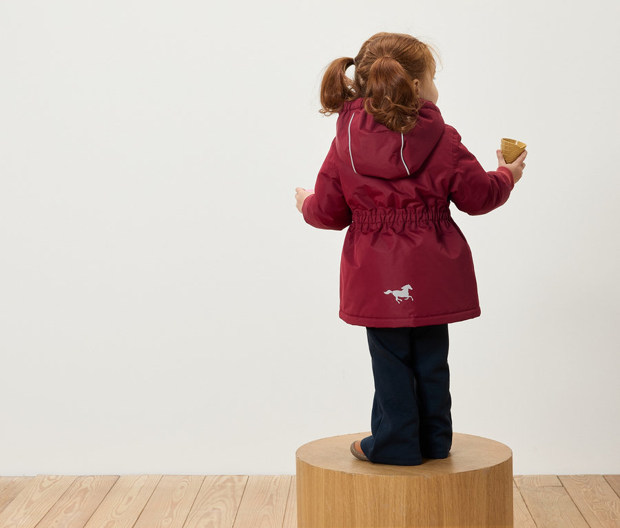 Ein rothaariges Mädchen mit Zöpfen steht mit dem Rücken zur Kamera auf einem Holzsockel, trägt eine bordeauxrote Jacke mit Kapuze und einem Pferdebild und hält eine Eistüte in der Hand.