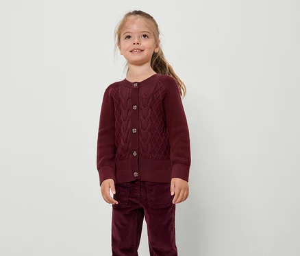 Mädchen in bordeauxrotem Pullover und Hose steht vor einem weißen Hintergrund.