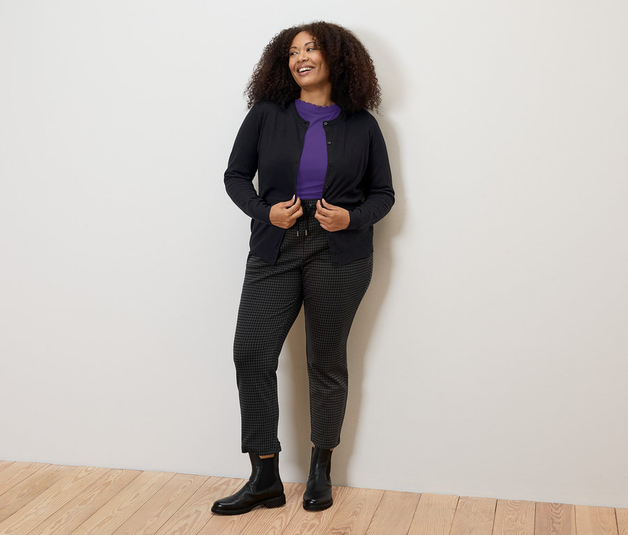 Frau trägt ein lila Shirt mit Wellenkante, eine dunkle Hose und eine Strickjacke.