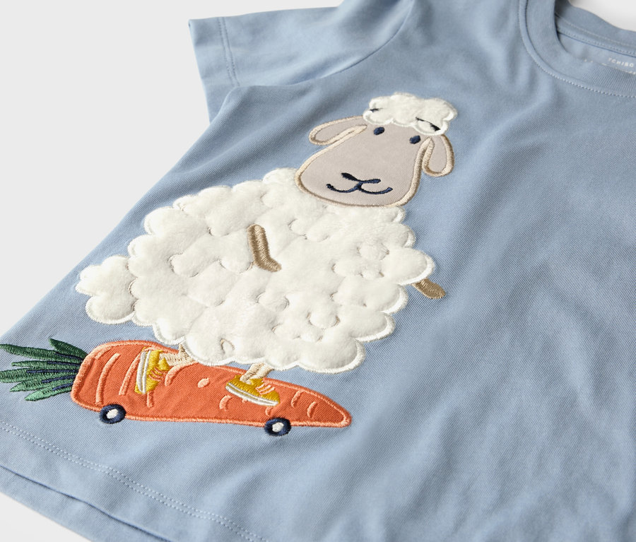 Hellblaues T-Shirt mit Applikation eines Schafs, das auf einer Karotte mit Rädern fährt.