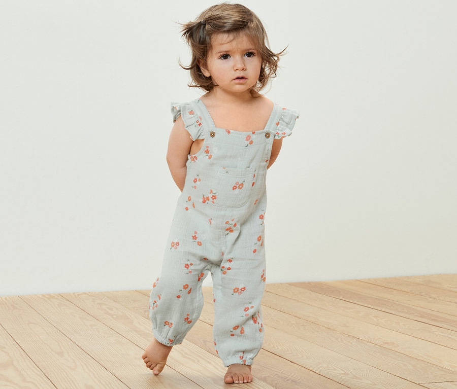 Kleinkind steht auf einem Holzboden und trägt einen Kinder-Musselin-Strampler mit Blumenmuster.