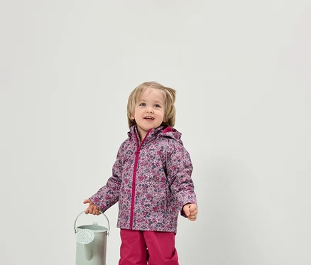 Kleinkind hält eine kleine grüne Gießkanne. Es trägt eine Jacke mit Blumenmuster und eine pinke Hose.