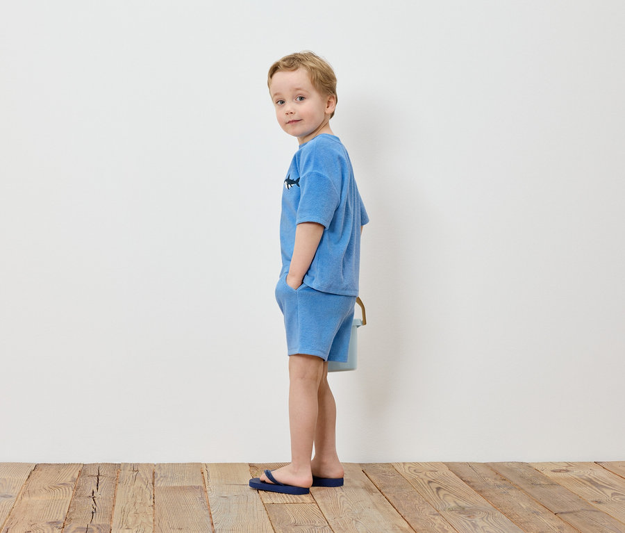Ein blonder Junge steht mit den Händen in den Taschen vor einer weißen Wand, gekleidet in ein hellblaues T-Shirt und Shorts, dazu blaue Flip-Flops.