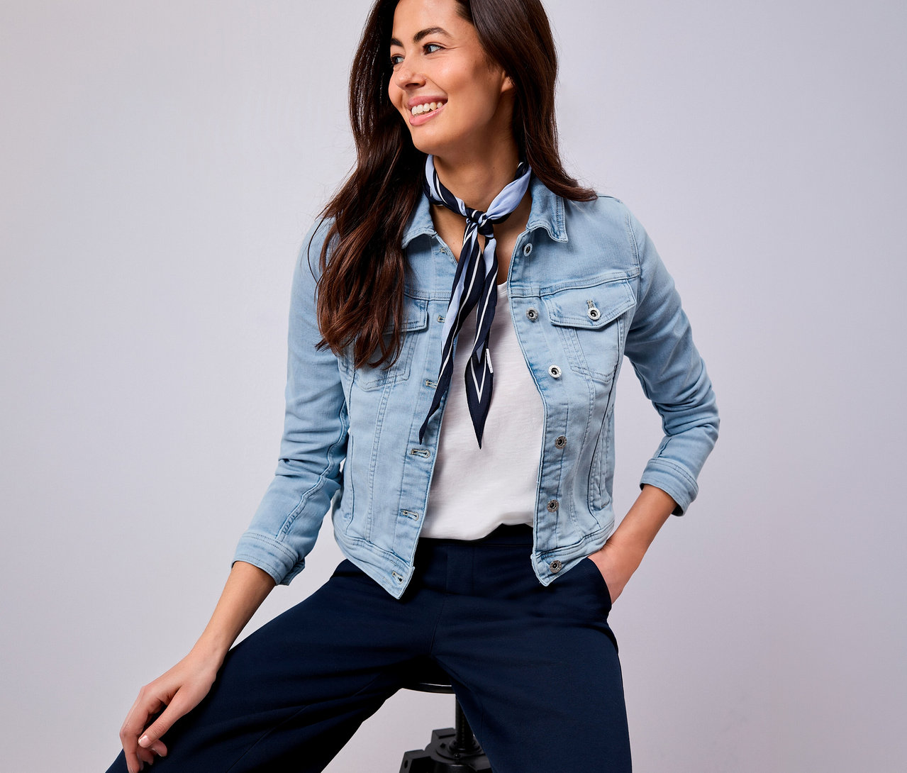 Lächelnde Frau mit blau-weiß gestreiftem Tuch, Jeansjacke und blauer Hose sitzt auf einem Hocker.