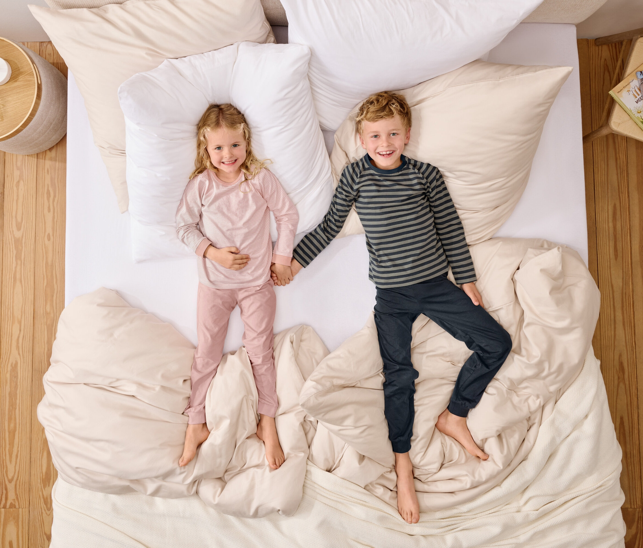 Draufsicht auf ein Mädchen in rosa Pyjamas und einen Jungen in gestreiftem Oberteil, die Händchen haltend auf einem Bett mit weißen Kissen liegen.