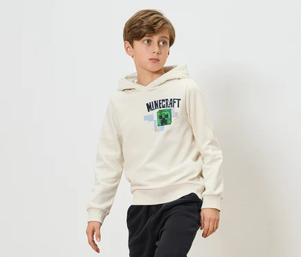 Ein Junge trägt einen cremefarbenen Minecraft-Kapuzenpullover und schwarze Jogginghosen vor einem weißen Hintergrund.