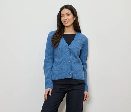 Frau mit dunklen Haaren trägt eine blaue Strickjacke mit drei Knöpfen über einem schwarzen Oberteil und einer dunklen Hose.
