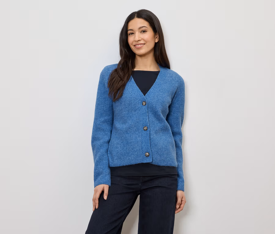 Frau mit dunklen Haaren trägt eine blaue Strickjacke mit drei Knöpfen über einem schwarzen Oberteil und einer dunklen Hose.