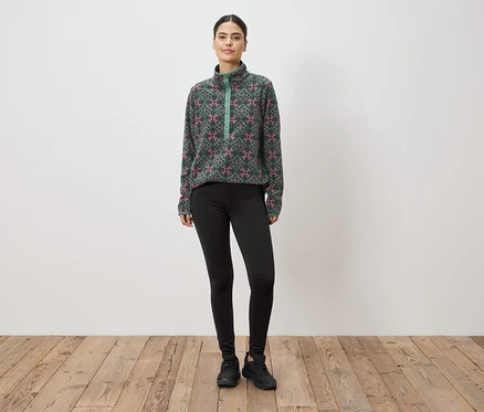 Eine Frau steht vor einer Wand und trägt ein grün-pink gemustertes Fleeceoberteil, Leggings und Turnschuhe.