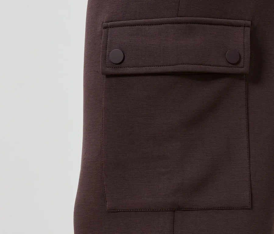 Detailansicht einer kastanienbraunen Hose mit aufgesetzter Tasche, die mit zwei runden Knöpfen geschlossen wird.