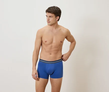 Mann ohne Hemd trägt blaue Boxershorts vor hellem Hintergrund.