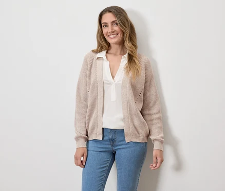 Frau mit beigem Cardigan, weißer Bluse und Jeans vor einer weißen Wand.