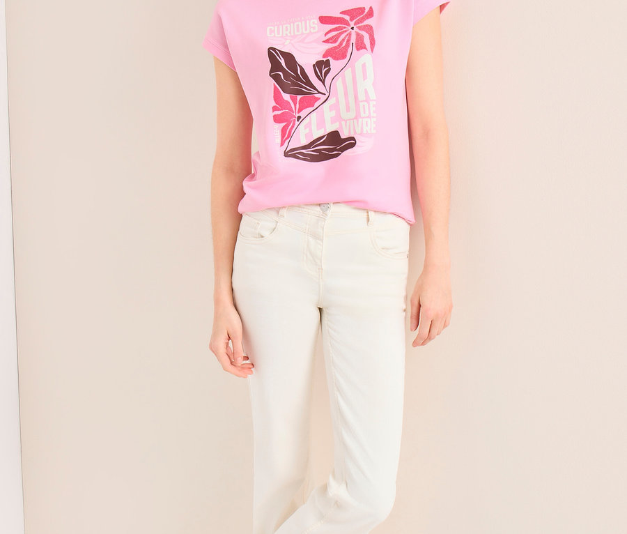 Eine lächelnde Frau mit beiger Kappe und hellen Jeans lehnt an einer Wand. Sie trägt ein pinkes CECIL Placement-Shirt mit floralem Aufdruck.