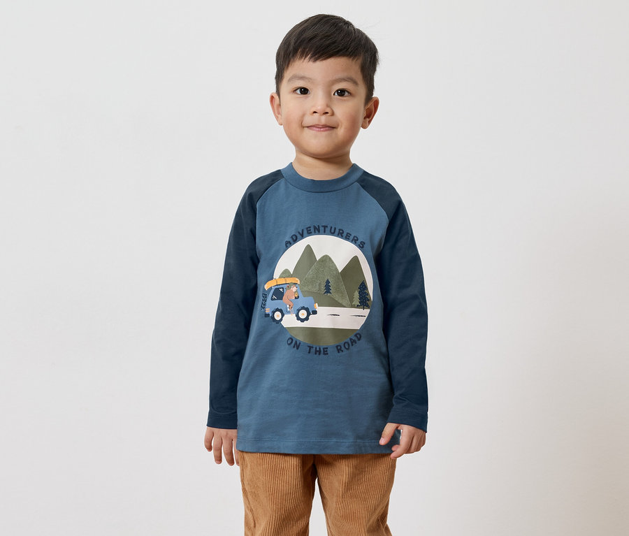 Junge mit blauem Langarmshirt mit Bären-Auto-Motiv und brauner Cordhose vor weißem Hintergrund.