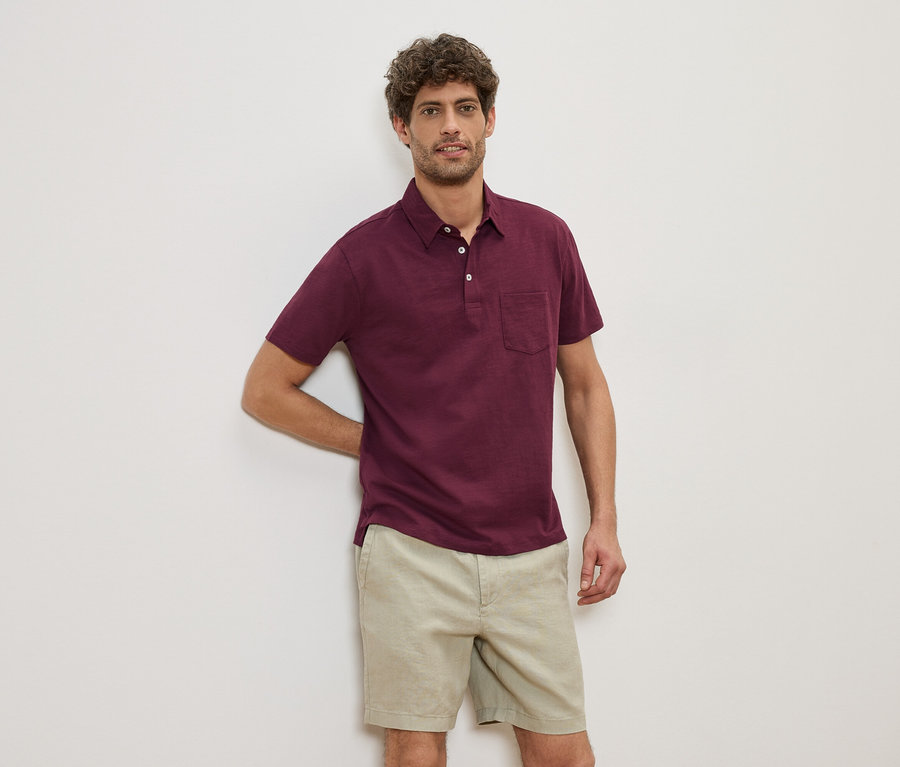 Ein Mann lehnt an einer Wand und trägt ein weinrotes Poloshirt und beige Shorts.