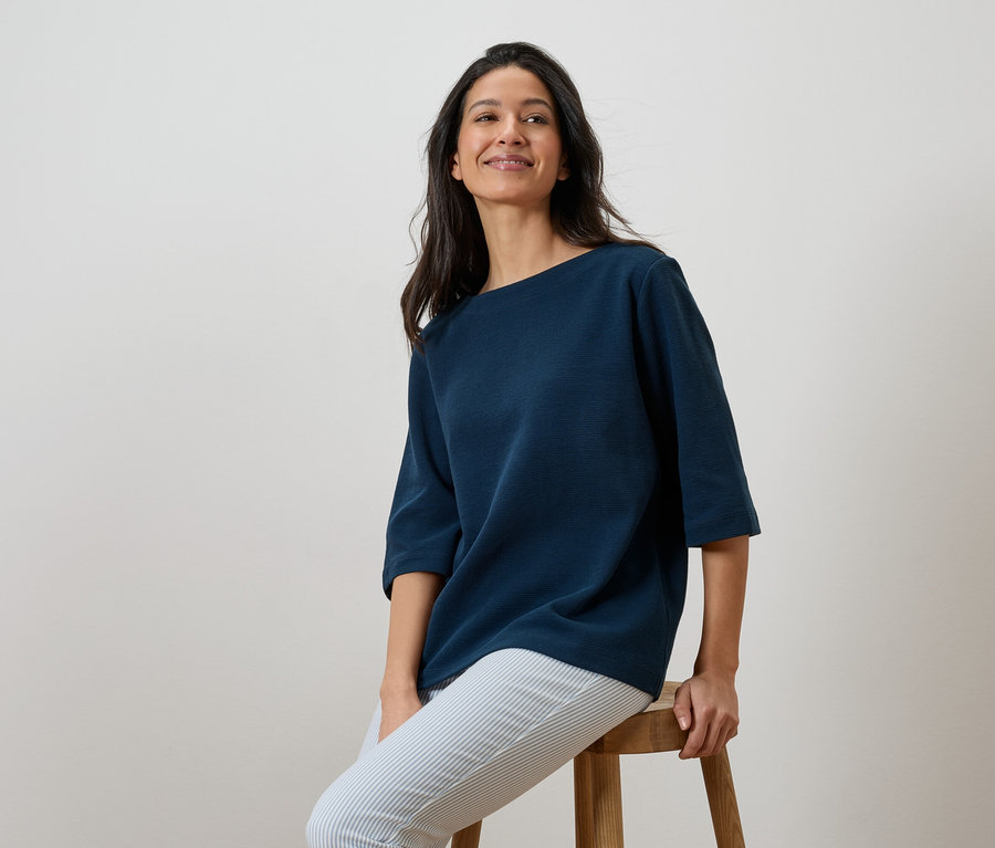 Frau sitzt auf einem Hocker und trägt ein marineblaues Shirt und gestreifte Hosen.