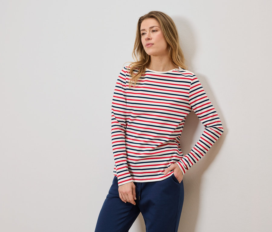 Frau mit gestreiftem Langarmshirt und blauer Jogginghose, eine Hand in der Hosentasche.