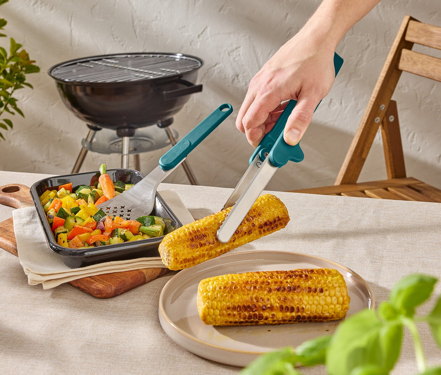Eine Person hält mit einer Grillzange einen Maiskolben über einen Teller. Auf dem Tisch stehen außerdem Gemüse und ein Grill.
