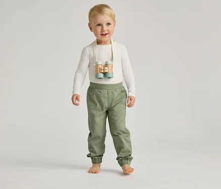 Ein barfüßiger Junge mit blauen Augen und blonden Haaren steht mit einer hellen Hose und einem hellen Oberteil vor einem grauen Hintergrund.
