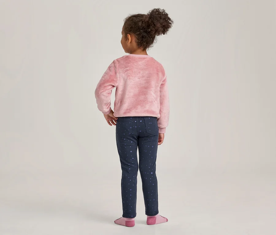 Junges Mädchen von hinten, trägt einen rosa flauschigen Pullover und dunkelblaue Leggings mit hellem Sternenmuster. Ihre lockigen Haare sind zu einem Dutt gebunden, die Hände sind in die Hüften gestemmt. Sie trägt rosa Socken.