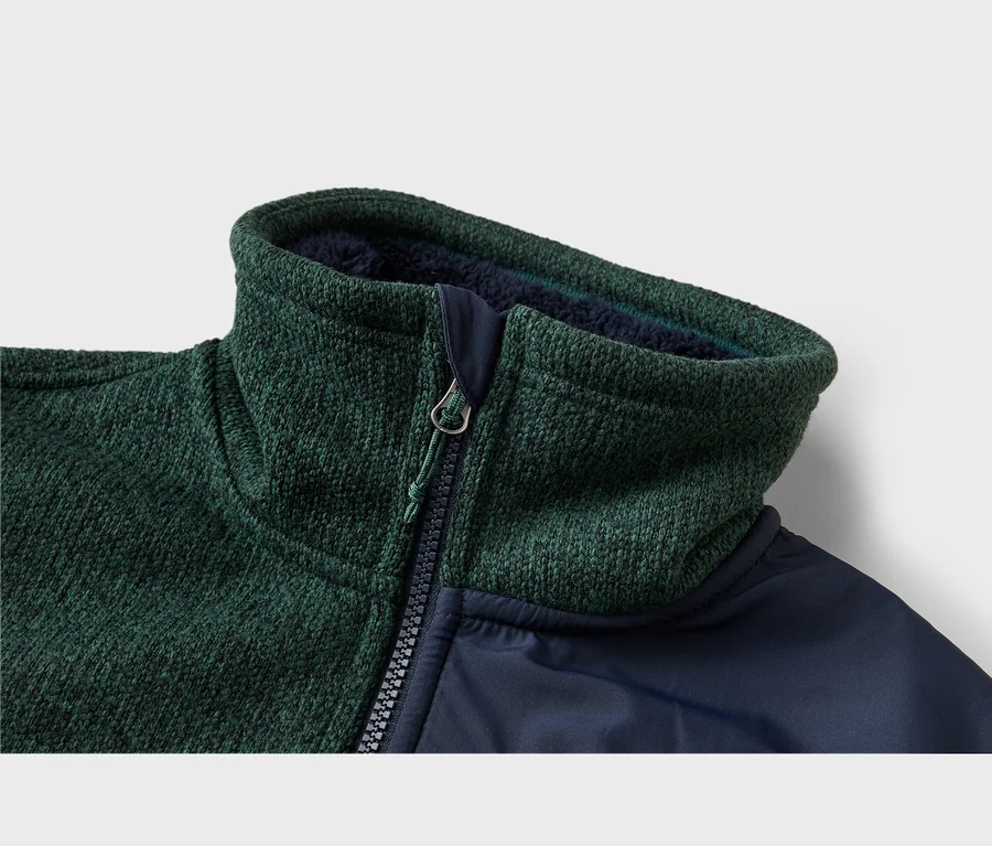 Nahaufnahme einer dunkelgrünen, strukturierten Jacke mit dunkelblauem, glattem Stoff. Hoher Kragen mit dunkelblauem Futter und schwarzem Reißverschluss mit silbernem Zipper und grünem Band.
