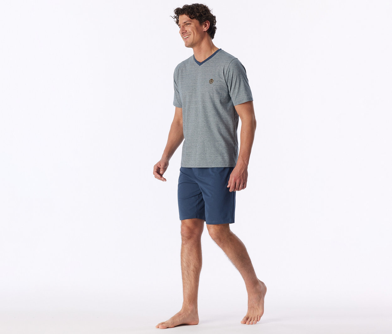 Ein lächelnder Mann mit lockigem braunem Haar geht barfuß und trägt ein graues V-Ausschnitt-T-Shirt mit kleinem Emblem und marineblauen Shorts.