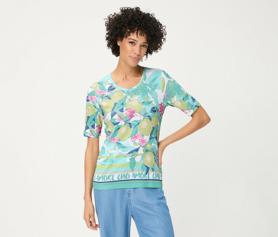 Frau mit lockigem Haar posiert in einem Olsen T-Shirt mit hellblauem Hintergrund, grünen Zitrusfrüchten, rosa Blüten und „AMORE CIAO“-Schriftzug am Saum, kombiniert mit hellblauer Hose.