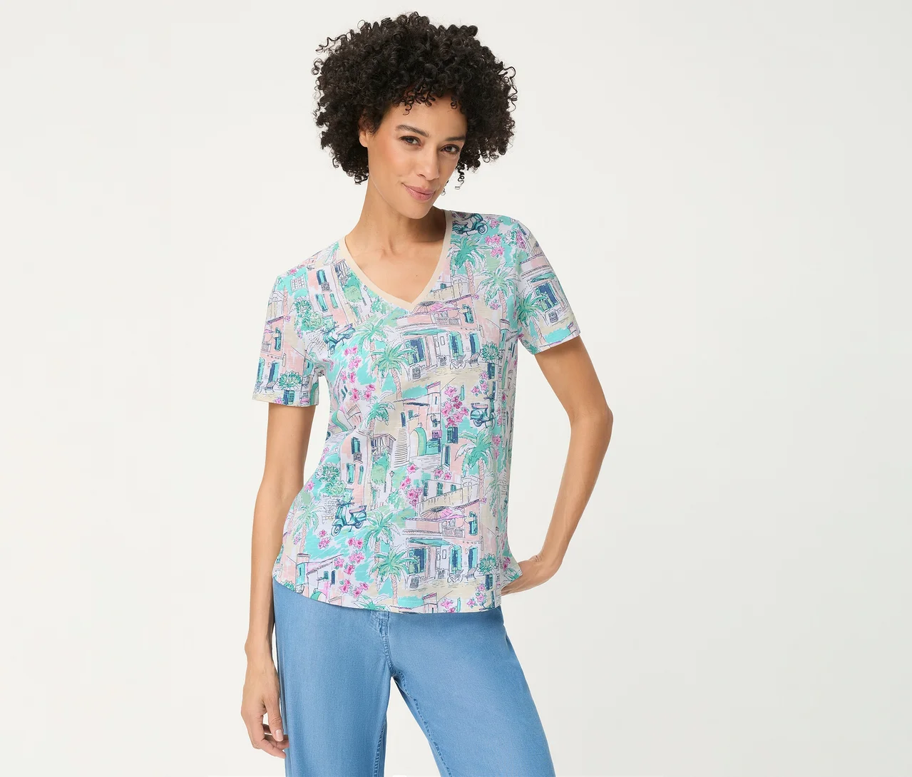 Frau mit Locken trägt ein Olsen T-Shirt mit pastellfarbenem Druck aus Gebäuden, Palmen und Blumen, dazu hellblaue Hosen, lächelt mit einer Hand an der Hüfte.