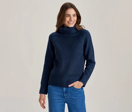Lächelnde Frau mit langen braunen Haaren, die einen dunkelblauen gerippten Rollkragenpullover und blaue Jeans trägt, eine Hand in der Tasche.