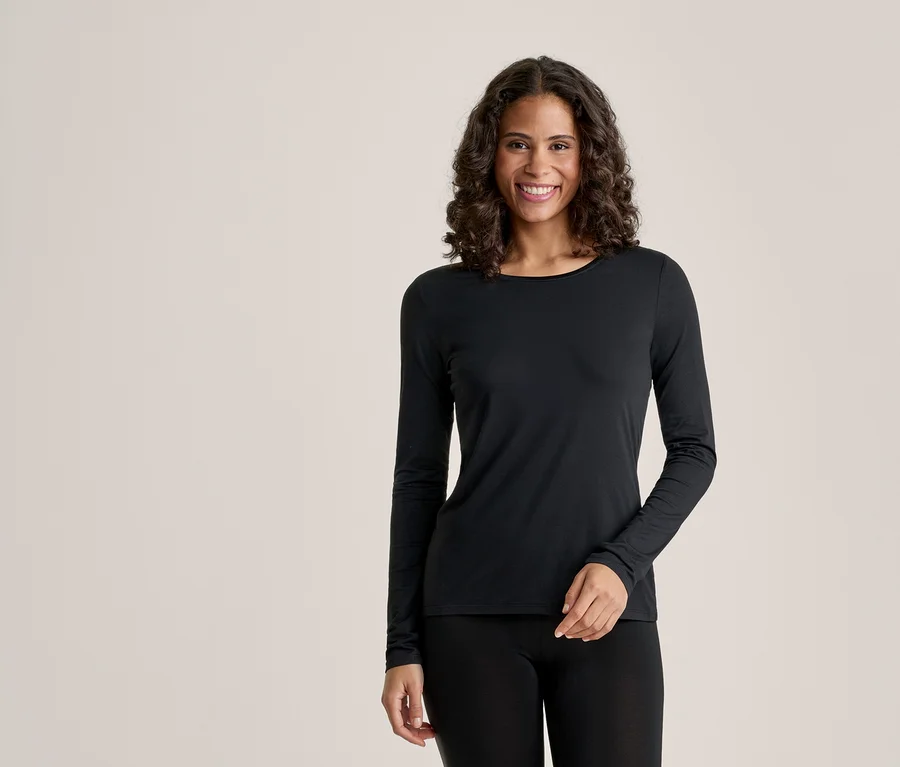 Eine lächelnde Frau mit dunklem, lockigem Haar trägt ein schwarzes Langarmshirt und schwarze Leggings vor einem hellen Hintergrund.
