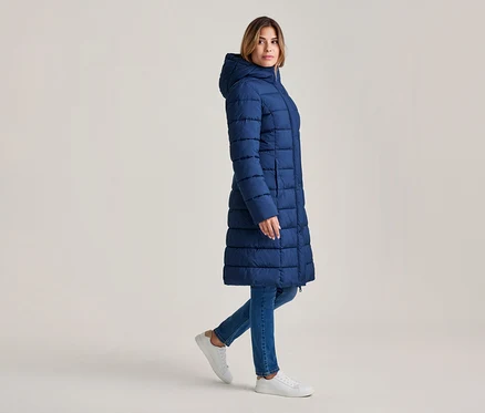 Frau geht, trägt einen langen, marineblauen Steppmantel mit Kapuze, blaue Jeans und weiße Sneaker.