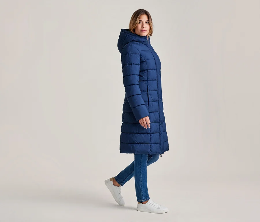 Frau geht, trägt einen langen, marineblauen Steppmantel mit Kapuze, blaue Jeans und weiße Sneaker.