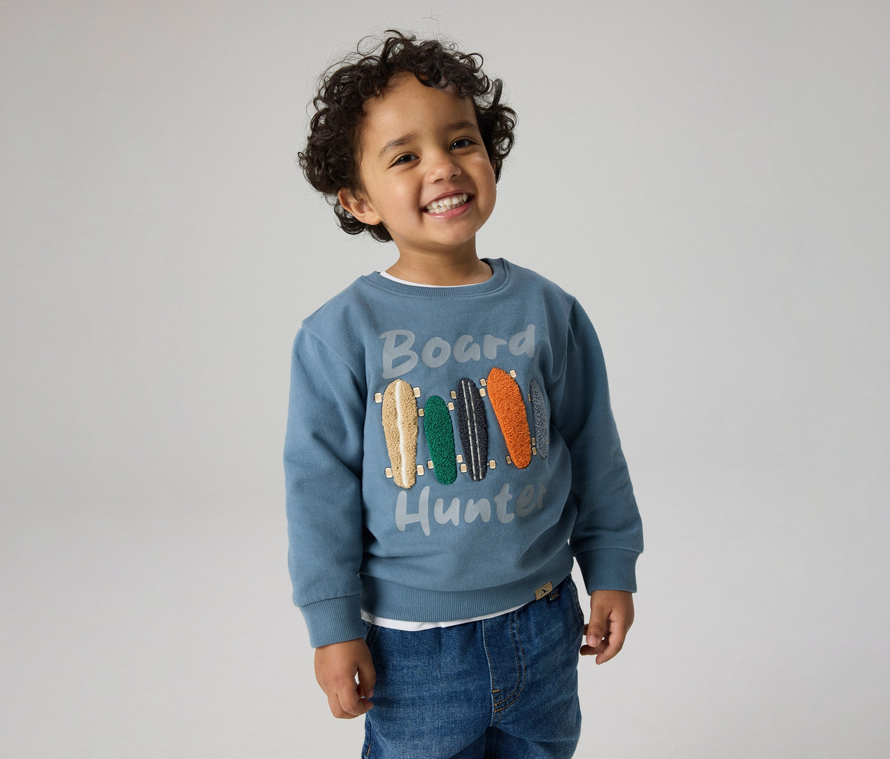 Lächelnder kleiner Junge mit lockigem Haar, der einen blauen Pullover mit "Board Hunter"-Schriftzug und Skateboard-Motiven sowie Jeans trägt.
