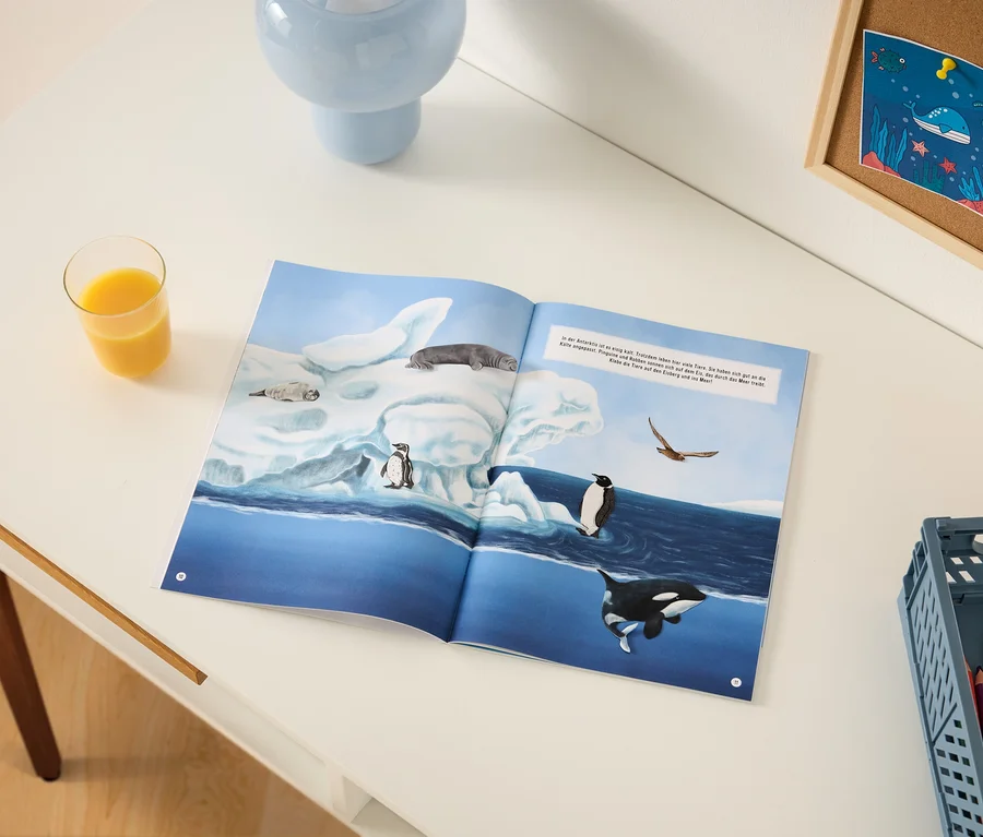 Ein aufgeschlagenes Kinderbuch auf einem weißen Schreibtisch zeigt eine Illustration von antarktischen Tieren wie Robben, Pinguinen und einem Orca auf Eisbergen und blauem Wasser. Ein Glas Orangensaft, eine hellblaue Vase, eine Korktafel mit einer Meeresbewohnerzeichnung und eine graue Kiste mit Buntstiften liegen ebenfalls auf dem Schreibtisch.