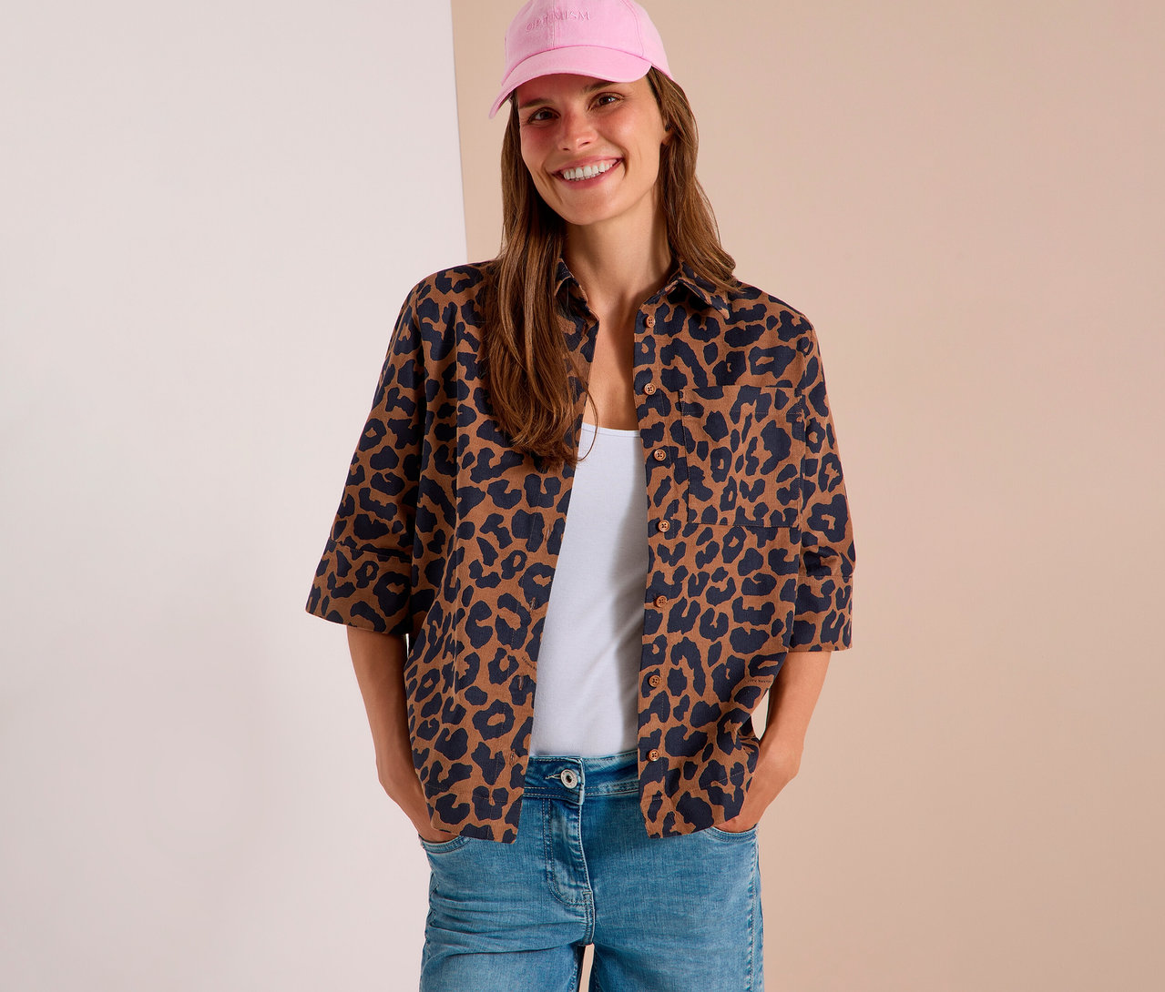 Lachende Frau mit pinker Baseballkappe, offener CECIL Hemdbluse mit Leopardenmuster über weißem Top und hellblauer Jeans, Hände in den Taschen.