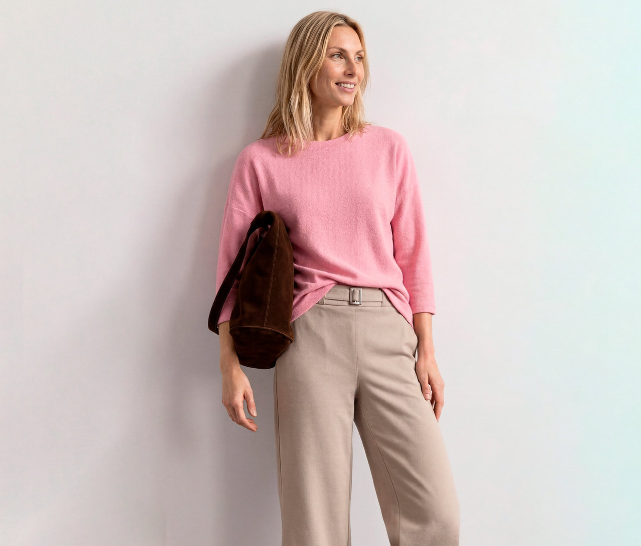 Lächelnde blonde Frau in einem pinken Street One 3/4-Arm-Shirt, beiger Hose und brauner Tasche, lehnt an einer weißen Wand.