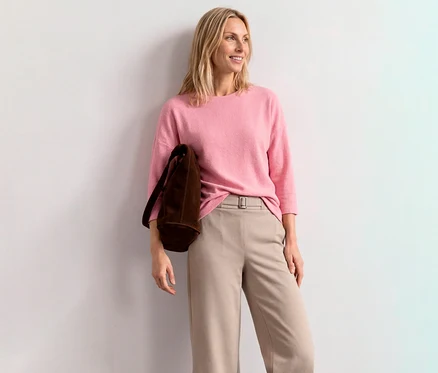 Lächelnde blonde Frau in einem pinken Street One 3/4-Arm-Shirt, beiger Hose und brauner Tasche, lehnt an einer weißen Wand.