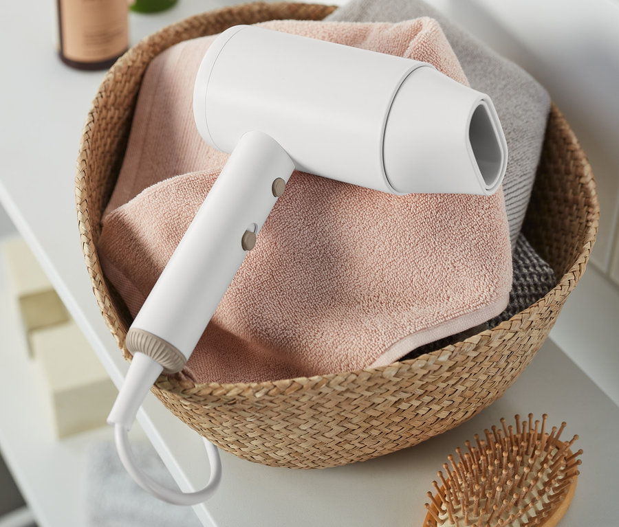 Weißer Haartrockner in einem Weidenkorb mit Handtüchern und einer Holzbürste auf einem weißen Regal.