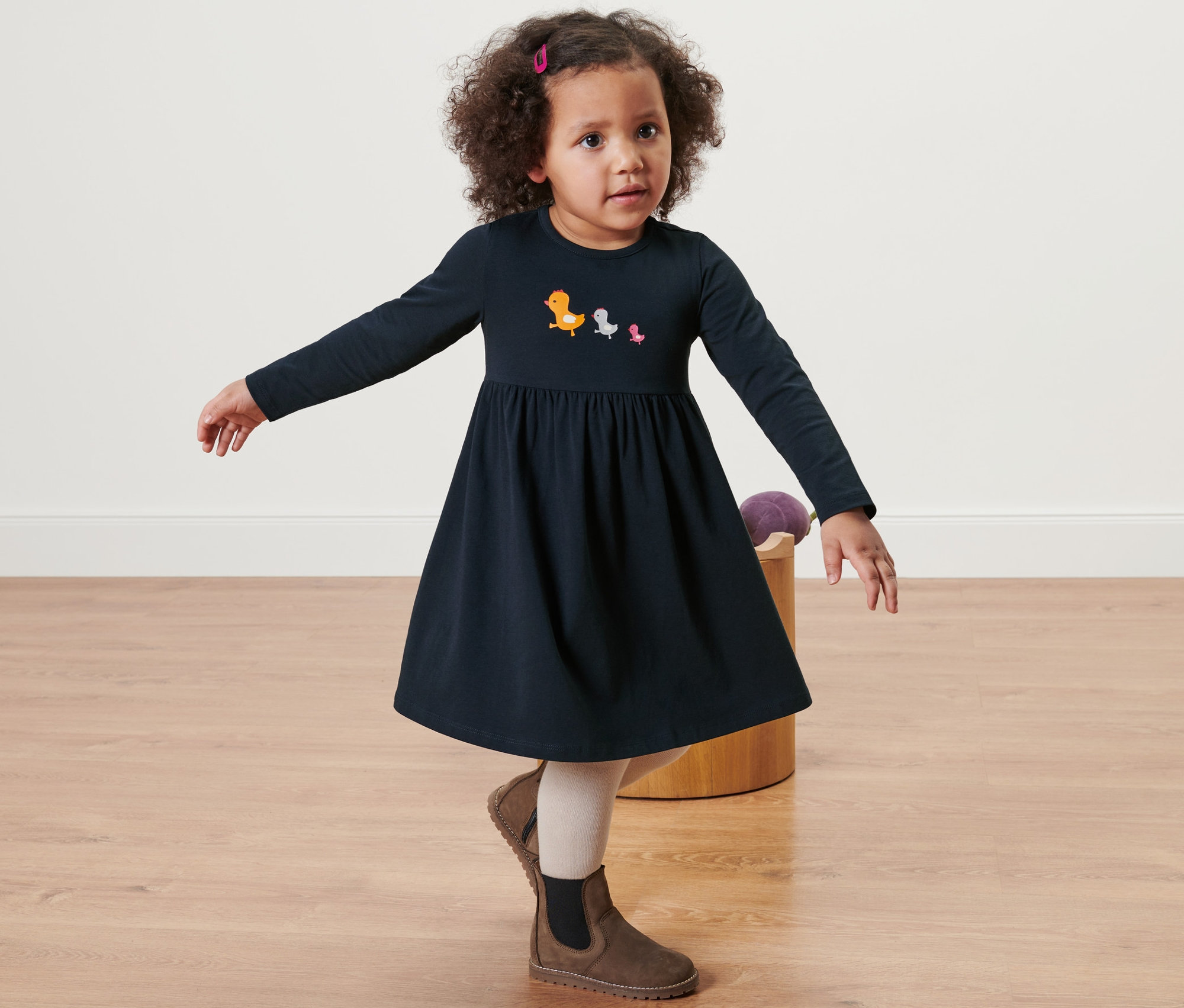 Mädchen posiert in einem blauen Baby Jerseykleid mit Langärmeln und Vogelmotiven, dazu braune Stiefel.